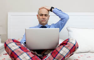 Man sitting up in bed, with a laptop and wearing a shirt and tie with pyjama trousers, looking unhappy.